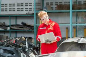 mecánico hombre inspeccionando coche partes valores en ordenador portátil computadora mientras trabajando en garaje depósito, joven mecánico con ordenador portátil en auto reparar tienda foto