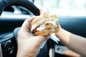 asiático mujer conductor sostener y comer hamburguesa en auto, peligroso y riesgo un accidente. foto