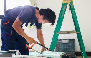 artesano trabajador aserradura un tubo, técnico hombre instalando un aire acondicionamiento en un cliente casa, joven reparador fijación aire acondicionador unidad, mantenimiento y reparando conceptos foto