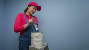 woman dressed in overalls and red t-shirt standing on a metal ladder taking the brush out of the jar of blue paint next to a blue wall photo