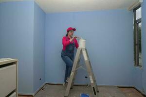 woman dressed in overalls and red t-shirt going up a metal ladder with a jar of paint next to a blue wall in an empty room photo