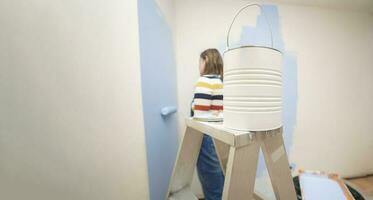 jar of paint on a ladder with a background of a standing woman dressed in overalls and striped blouse, seen from behind, painting a white wall with a roller with blue paint photo