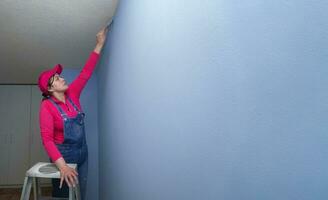 woman dressed in overalls and a red t-shirt on a metal ladder, with a brush in her hand finishing painting a blue wall in an empty room photo