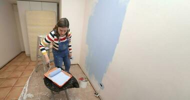 mujer vestido en mono y a rayas blusa, visto desde el frente, inmersión un rodillo en el bandeja con azul pintar, dentro un vacío habitación con blanco paredes medio pintado con azul pintar foto