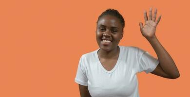 retrato de joven africano mujer mirando a cámara y ondulación mano con grande sonrisa en contra naranja antecedentes foto