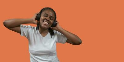 Portrait of african young woman listening to music on headphones with toothy smile, holding headphones with hands against orange colored background photo