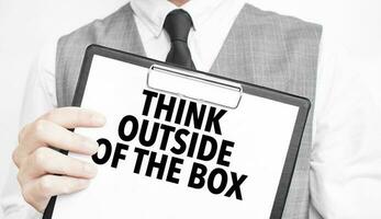 think outside of the box inscription on a notebook in the hands of a businessman on a grey background, a man points with a finger to the text photo