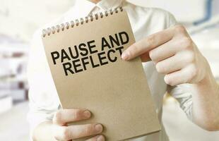 Text PAUSE AND REFLECT on brown paper notepad in businessman hands in office. Business concept photo