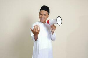 Portrait of attractive Asian muslim man in white shirt with skullcap speaking louder using megaphone, promoting product. Advertising concept. Isolated image on grey background photo