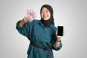 Portrait of excited Asian muslim woman with hijab, showing one hundred thousand rupiah while showing blank screen mockup mobile phone. Financial and savings concept. Isolated image on white background photo