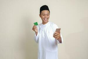 Portrait of attractive Asian muslim man in white shirt with skullcap holding a mobile phone and presenting credit card. Isolated image on gray background photo