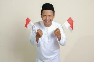 Portrait of attractive Asian muslim man in white shirt with skullcap holding indonesia flag while raising his fist, celebrating success. Isolated image on gray background photo