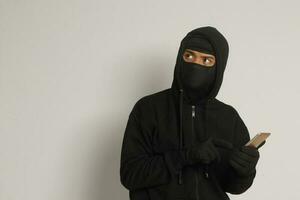 Portrait of mysterious man wearing black hoodie and mask doing hacking activity on mobile phone, hacker holding a smartphone. Isolated image on gray background photo