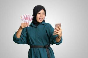 Portrait of excited Asian hijab woman in casual outfit showing one hundred thousand rupiah while holding a mobile phone. Financial and savings concept. Isolated image on white background photo