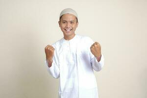 Portrait of attractive Asian muslim man in white shirt raising his fist, celebrating success. Isolated image on gray background photo