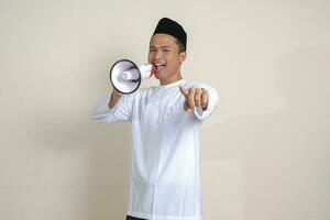 Portrait of attractive Asian muslim man in white shirt with skullcap speaking louder using megaphone, promoting product. Advertising concept. Isolated image on grey background photo