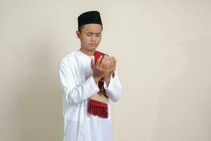 retrato de atractivo asiático musulmán hombre en blanco camisa con casquete Orando formalmente con su manos aumentó. aislado imagen en gris antecedentes foto