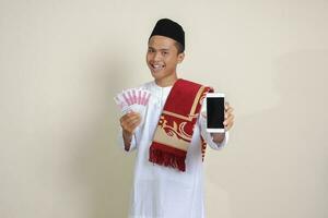 Portrait of attractive Asian muslim man in white shirt with skullcap showing one hundred thousand rupiah while showing blank screen mobile phone. Financial and shopping concept. Isolated image on gray photo