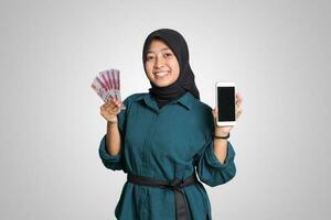 Portrait of excited Asian muslim woman with hijab, showing one hundred thousand rupiah while showing blank screen mockup mobile phone. Financial and savings concept. Isolated image on white background photo