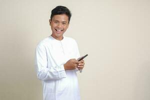 Portrait of attractive Asian muslim man in white shirt holding mobile phone with smiling expression on face. Advertising concept. Isolated image on gray photo