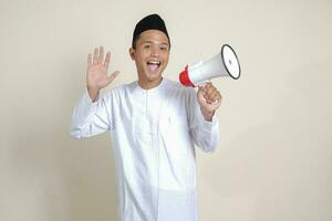 Portrait of attractive Asian muslim man in white shirt with skullcap speaking louder using megaphone, promoting product. Advertising concept. Isolated image on grey background photo