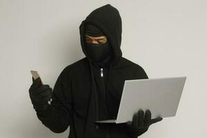 Portrait of mysterious man wearing black hoodie and mask doing hacking activity on laptop, hacker holding a mobile phone. Cyber security concept. Isolated image on gray background photo