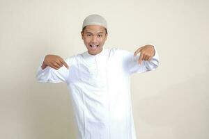 Portrait of attractive Asian muslim man in white shirt with skullcap showing product and pointing with his hand and finger to the side. Advertising concept. Isolated image on gray background photo
