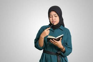 Portrait of excited Asian muslim woman with hijab writing on note book and thinking with pen on chin. Advertising concept. Isolated image on white background photo