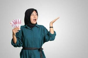 Portrait of excited Asian hijab woman in casual outfit showing and pointing one hundred thousand rupiah. Financial and savings concept. Isolated image on white background photo