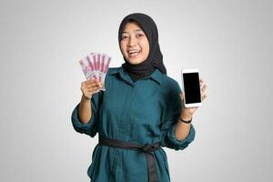 Portrait of excited Asian muslim woman with hijab, showing one hundred thousand rupiah while showing blank screen mockup mobile phone. Financial and savings concept. Isolated image on white background photo