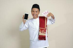 retrato de atractivo asiático musulmán hombre en blanco camisa con casquete demostración uno cien mil rupia mientras demostración blanco pantalla móvil teléfono. financiero y compras concepto. aislado imagen en gris foto