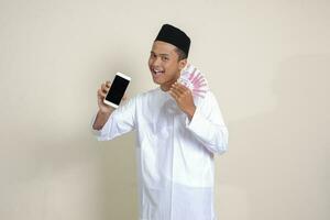 Portrait of attractive Asian muslim man in white shirt with skullcap showing one hundred thousand rupiah while showing blank screen mobile phone. Financial and shopping concept. Isolated image on gray photo