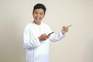 Portrait of attractive Asian muslim man in white shirt holding mobile phone with smiling expression on face while pointing finger to the side. Advertising concept. Isolated image on gray background photo