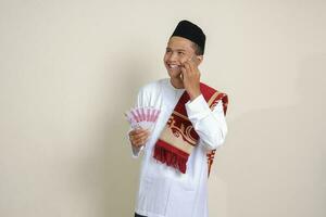 Portrait of attractive Asian muslim man in white shirt showing one hundred thousand rupiah while using mobile phone. Financial and savings concept. Isolated image on gray background photo