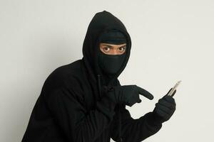 Portrait of mysterious man wearing black hoodie and mask doing hacking activity on mobile phone, hacker holding a smartphone. Isolated image on gray background photo