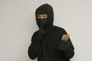 Portrait of mysterious man wearing black hoodie and mask doing hacking activity on mobile phone, hacker holding a smartphone. Isolated image on gray background photo