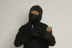 Portrait of mysterious man wearing black hoodie and mask doing hacking activity on mobile phone, hacker holding a smartphone. Isolated image on gray background photo