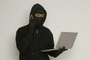 Portrait of mysterious man wearing black hoodie and mask doing hacking activity on laptop, hacker holding a mobile phone. Cyber security concept. Isolated image on gray background photo