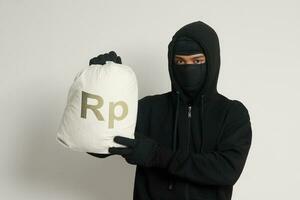 misterioso ladrón ladrón hombre vistiendo negro capucha y máscara que lleva un bolso lleno de rupia dinero. aislado imagen en gris antecedentes foto