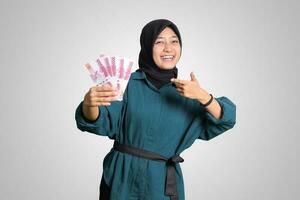 Portrait of excited Asian hijab woman in casual outfit showing and pointing one hundred thousand rupiah. Financial and savings concept. Isolated image on white background photo