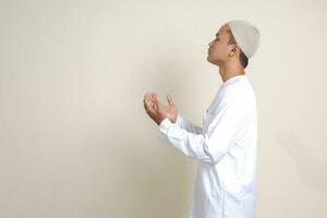 Portrait of attractive Asian muslim man in white shirt with skullcap praying earnestly with his hands raised. Isolated image on gray background photo