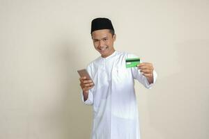 Portrait of attractive Asian muslim man in white shirt with skullcap holding a mobile phone and presenting credit card. Isolated image on gray background photo