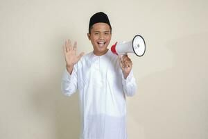 Portrait of attractive Asian muslim man in white shirt with skullcap speaking louder using megaphone, promoting product. Advertising concept. Isolated image on grey background photo