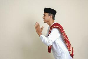 Portrait of attractive Asian muslim man in white shirt with skullcap showing apologize and welcome hand gesture. Isolated image on gray background photo
