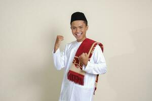 Portrait of attractive Asian muslim man in white shirt raising his fist, celebrating success. Isolated image on gray background photo