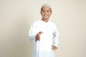 Portrait of attractive Asian muslim man in white shirt reaching out his hand for a handshake, welcoming someone. Isolated image on gray background photo