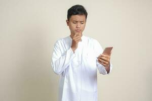 Portrait of attractive Asian muslim man in white shirt thinking about question with hand on chin while holding a mobile phone. Isolated image on gray background photo