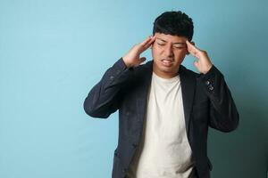 Portrait of young Asian business man in casual suit with dizzy expression while holding head with both hands in stress. Isolated image on blue background photo