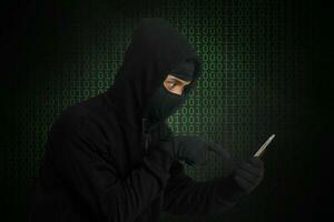 Portrait of mysterious man wearing black hoodie and mask doing hacking activity on mobile phone, hacker holding a smartphone. Isolated image on gray background photo