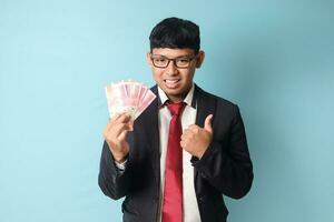 Portrait of happy young Asian business man in casual suit holding thousand rupiahs while making thumbs up. Isolated image on blue background photo